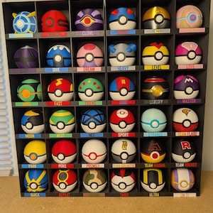 May include: A black grid display shelf containing thirty spherical containers, each featuring unique color patterns and designs. Each compartment includes a small label below the sphere with text identifying the specific type of ball, such as Poke, Great, Ultra, Master, and various team-themed designs.