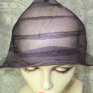 May include: A dark purple mesh hat with a brim and a conical crown. The hat has horizontal bands of stitching and is displayed on a mannequin head with closed eyes and red lipstick. The background is a lace curtain.