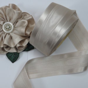 May include: A roll of beige satin ribbon with a subtle striped texture. A beige fabric flower with a pearl-centered button and green leaves is also visible. The ribbon is ideal for crafts, gift wrapping, or decorative purposes.
