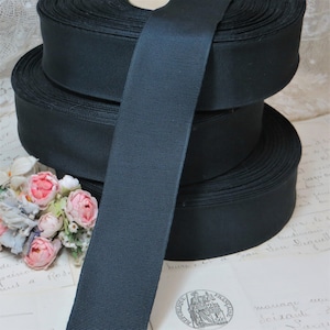 May include: A stack of black grosgrain ribbon rolls. The ribbon has a ribbed texture and is unspooled, revealing the width of the material. The image also shows a small bouquet of pink and white flowers.