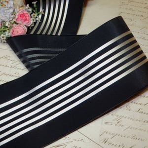 May include: Black satin ribbon with silver and white stripes. The ribbon is folded over itself, showing the pattern. The ribbon is on a vintage paper background with floral accents.