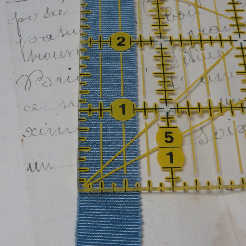 May include: A close-up of a blue fabric measuring tape with yellow markings. The tape is marked with numbers 1, 2, and 5. The tape is against a white background.