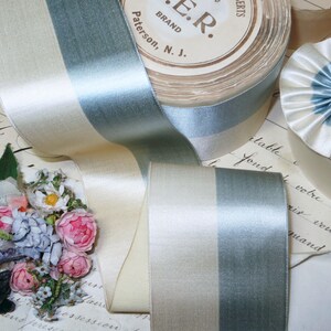 May include: A roll of satin ribbon in cream and pale blue, with a matching ribbon unfurled. The ribbon has a subtle sheen and is layered over vintage paper with floral accents. The label on the roll reads "E.R. BRAND Paterson, N.J."