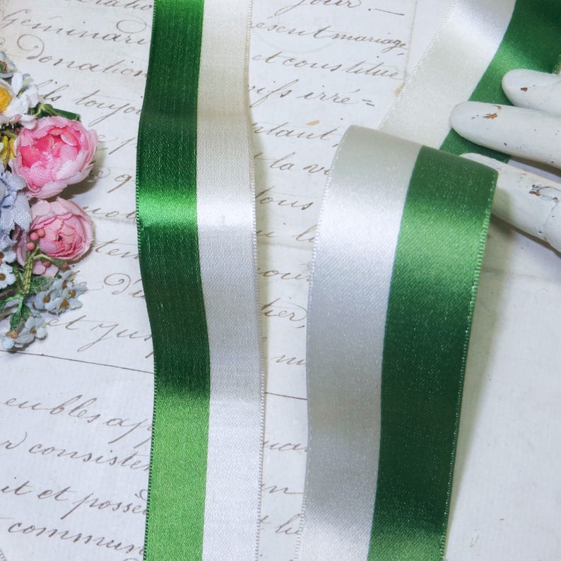 May include: Close-up of green and white satin ribbon. The ribbon is divided into three vertical stripes: green, white, and green. The ribbon is smooth and shiny, with a slight sheen. The background is a vintage paper with faded script.