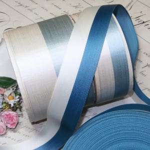 May include: Spools of satin ribbon in various shades of blue, white, and cream. The ribbons are arranged in a striped pattern, with a solid blue ribbon unspooled at the bottom. The ribbons are likely used for crafting or gift wrapping.