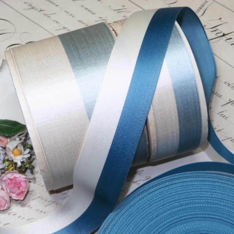 May include: Spools of satin ribbon in various shades of blue, white, and cream. The ribbons are arranged in a striped pattern, with a solid blue ribbon unspooled at the bottom. The ribbons are likely used for crafting or gift wrapping.