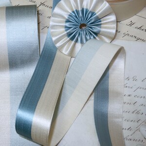 May include: Close-up of various ribbons and a rosette in shades of blue and cream. The ribbons have a satin finish and are striped with different widths of blue and cream. A circular rosette with a blue center and cream outer ring is also visible.