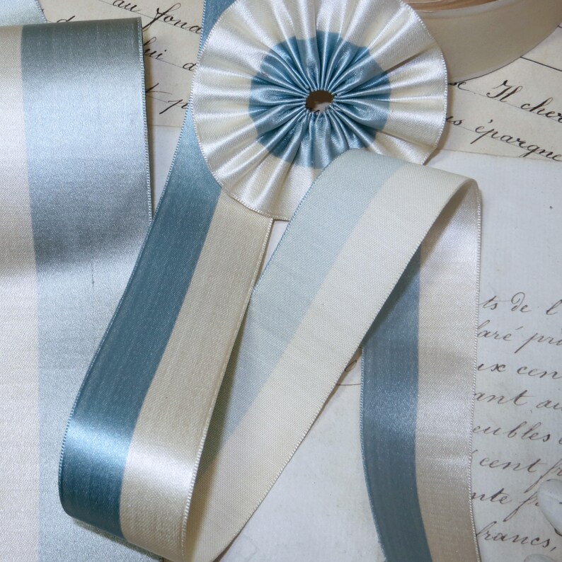 May include: Close-up of various ribbons and a rosette in shades of blue and cream. The ribbons have a satin finish and are striped with different widths of blue and cream. A circular rosette with a blue center and cream outer ring is also visible.
