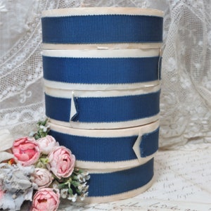 May include: A stack of spools of blue and white striped ribbon. The ribbon is a dark blue colour and has a white stripe running down the centre. The spools are stacked on top of each other and are sitting on a white surface.