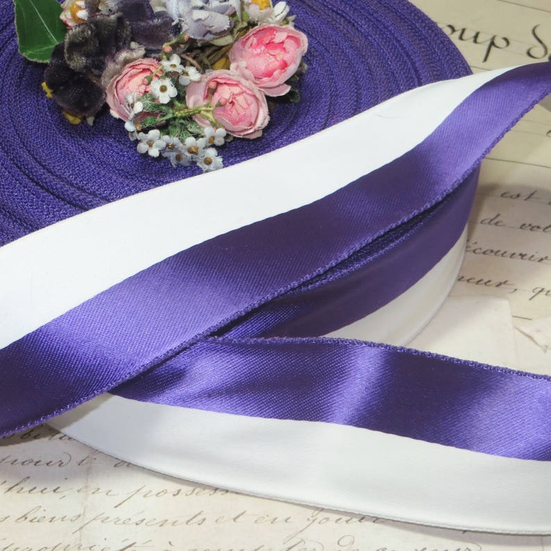 May include: A roll of purple and white satin ribbon with a floral arrangement on top. The ribbon is a deep purple with a white stripe down the center. The flowers are pink and white.