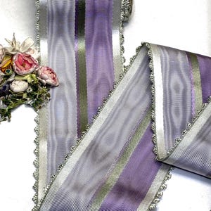 May include: A roll of decorative ribbon with a striped pattern in shades of lavender, silver, and olive green. The ribbon has a scalloped edge with silver trim, suitable for crafts and embellishments.