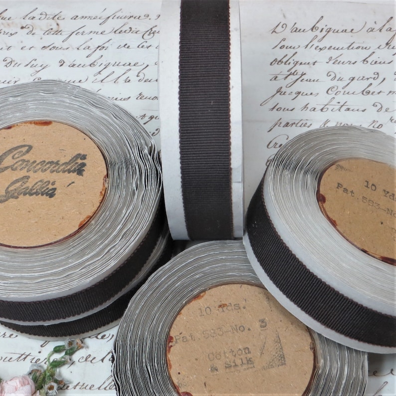 May include: Close-up of spools of brown grosgrain ribbon with white backing. The spools are labelled with "Concordia Gallia" and "Pat. 583-No. 3 Cotton & Silk".