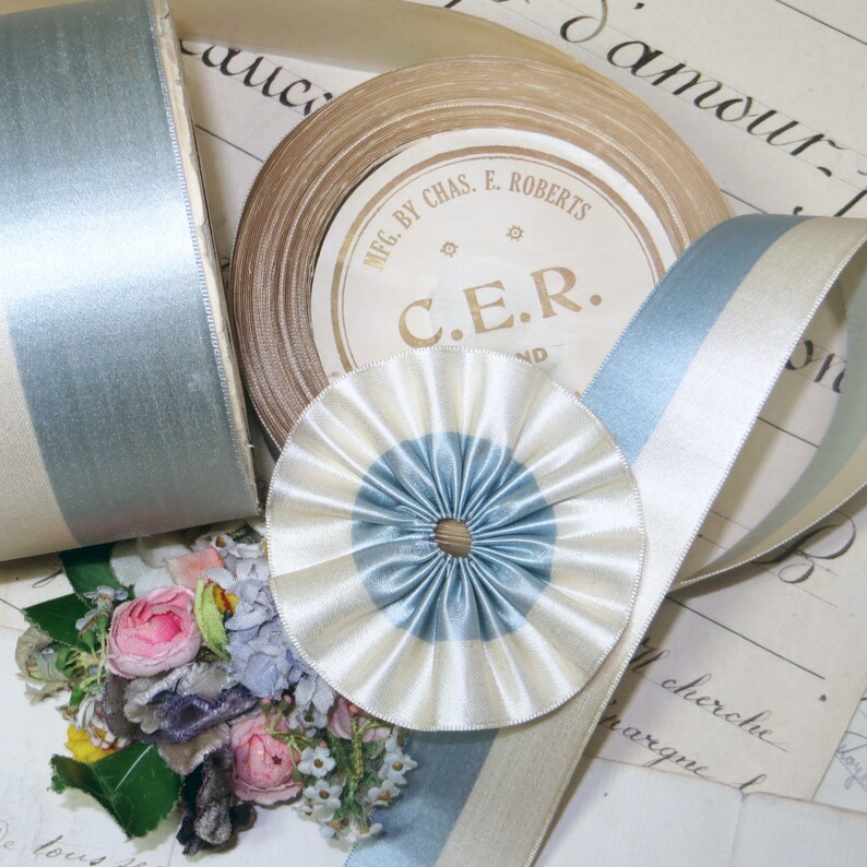 May include: An assortment of vintage ribbons in shades of blue, cream, and tan. A spool of tan ribbon with a label that reads "MFG. BY CHAS. E. ROBERTS C.E.R. ND" is visible. A rosette and a roll of ribbon are also present, along with a small bouquet.