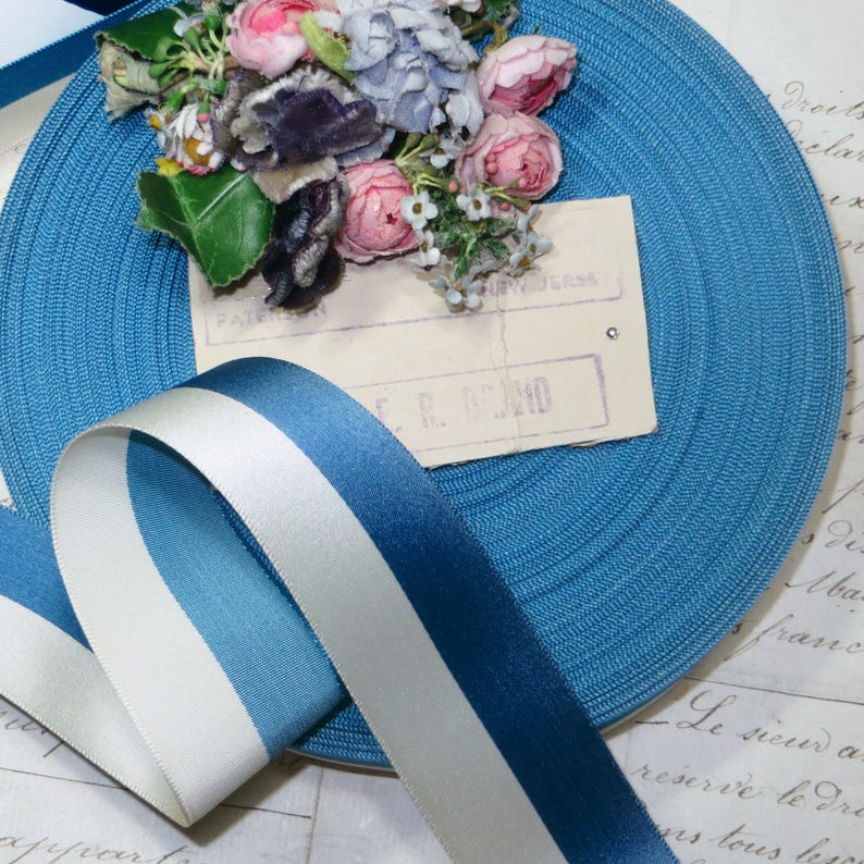 May include: A roll of turquoise blue ribbon with a textured surface, and a striped ribbon in shades of blue and white. A small bouquet of pink and purple flowers rests on a vintage paper. The background features script.
