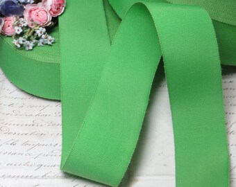 Puede incluir: Cinta de grogrén verde brillante, desenrollada sobre un fondo de papel vintage. La cinta es de un color sólido y vibrante con una textura acanalada. Un pequeño arreglo de flores rosas y azules se encuentra en el borde del rollo de cinta.