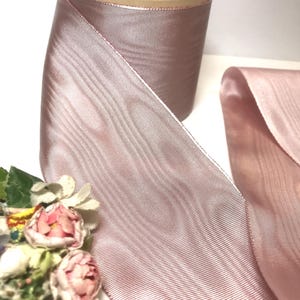 May include: A roll of dusty rose-colored ribbon with a subtle sheen. The ribbon is unwound, revealing its texture and color. The spool is made of cardboard. A small bouquet of pink and white flowers is in the foreground.