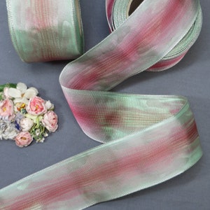 May include: A roll of shimmering ribbon in shades of green and pink. The ribbon has a watercolor effect, with the colors blending together. A small bundle of pink and white flowers is in the background. The ribbon is approximately 1 inch wide.
