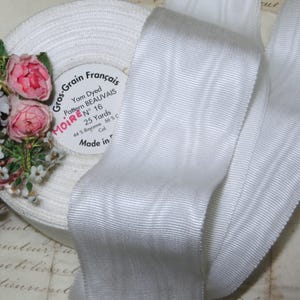 May include: A spool of white grosgrain ribbon with a textured surface, labeled "Gros-Grain Français." The label indicates "Yam Dyed" and "Pattern BEAUVAIS." The ribbon is 25 yards long. Pink and white floral accents are visible.
