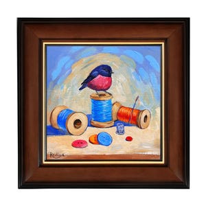 Puede incluir: Una pintura enmarcada de un pequeño pájaro azul y rosa posado en un carrete de hilo azul. Hay otros dos carretes de hilo, uno rojo y otro azul, y varios botones esparcidos alrededor de los carretes. La pintura está firmada "Redive" en la esquina inferior izquierda.