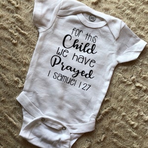 Op de afbeelding: Witte baby romper met zwarte tekst die "Voor dit kind hebben we gebeden 1 Samuel 127" luidt.
