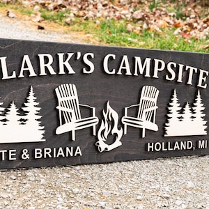 Puede incluir: Un letrero de madera marr&oacute;n oscuro con letras blancas que dice "Clark's Campsite" con dos siluetas blancas de sillas frente a una fogata y el texto "Nate & Briana" y "Holland, MI".