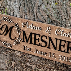 May include: A wooden sign with the text "Adam & Claire Mesner Est. June 15, 2019" in white lettering. A large "M" is in a wreath of leaves.