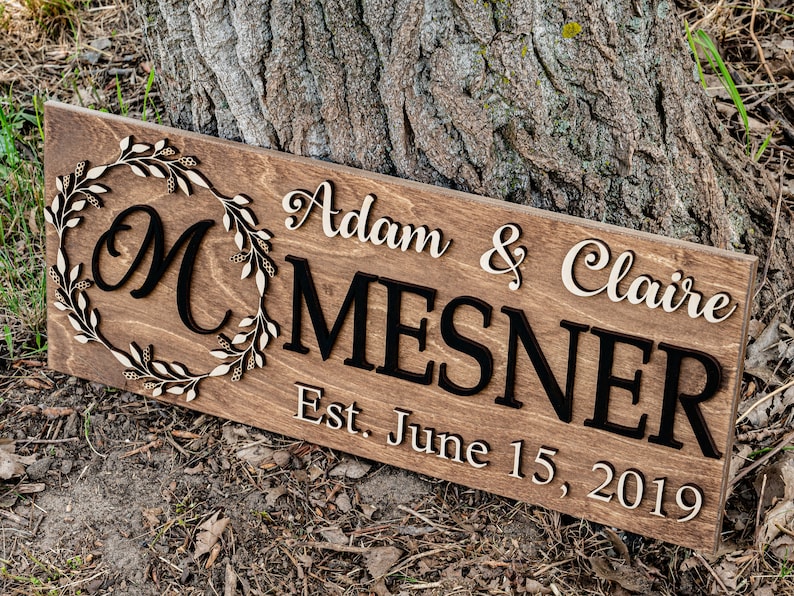 May include: A wooden sign with the text "Adam & Claire Mesner Est. June 15, 2019" in white lettering. A large "M" is in a wreath of leaves.