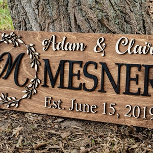 May include: A wooden sign with a wreath and the letters "M" in a circle. The text "Adam & Claire MESNER Est. June 15, 2019" is on the sign.