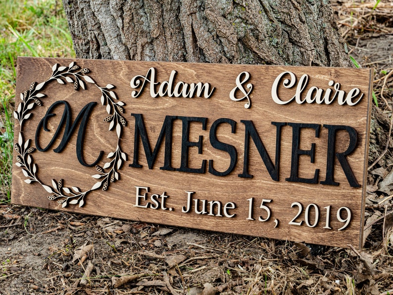 May include: A wooden sign with a wreath and the letters "M" in a circle. The text "Adam & Claire MESNER Est. June 15, 2019" is on the sign.