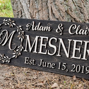 May include: A dark brown wooden sign with the text "Adam & Claire Mesner Est. June 15, 2019" in white lettering. A decorative wreath with a white "M" in the center is on the left side of the sign.