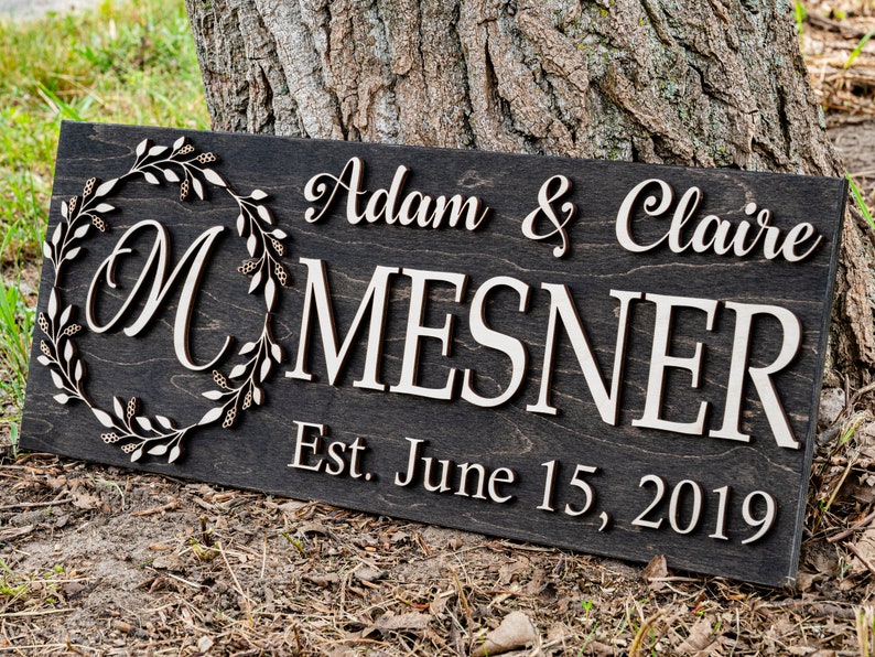 May include: A dark brown wooden sign with the text "Adam & Claire Mesner Est. June 15, 2019" in white lettering. A decorative wreath with a white "M" in the center is on the left side of the sign.