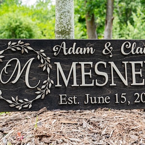 May include: A dark brown wooden sign with the text "Adam & Claire MESNER Est. June 15, 2019" in white lettering. A decorative wreath with a white "M" in the center is on the left side of the sign.