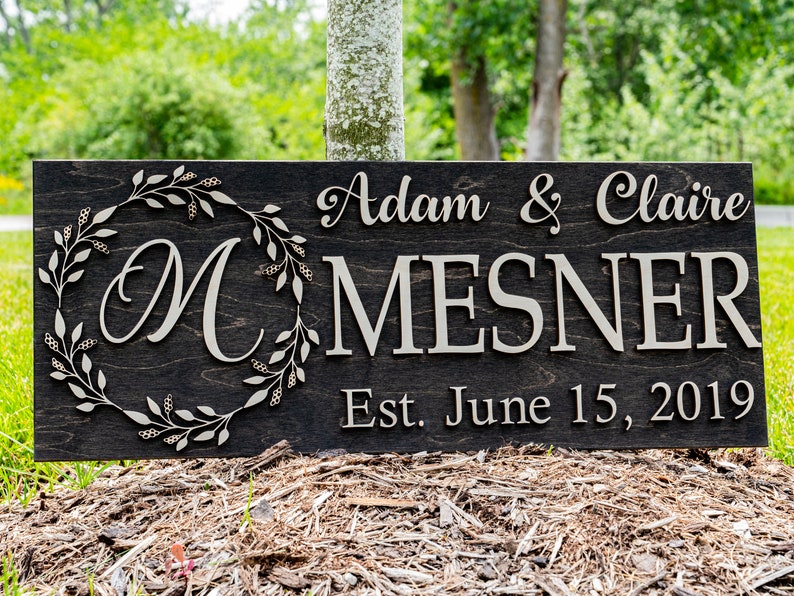 May include: A dark brown wooden sign with the text "Adam & Claire MESNER Est. June 15, 2019" in white lettering. A decorative wreath with a white "M" in the center is on the left side of the sign.