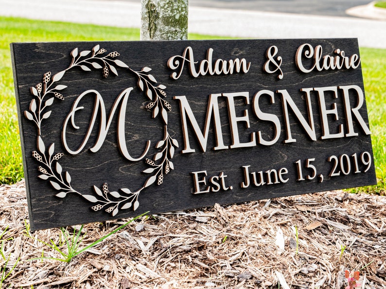 May include: A dark wood sign with the text "Adam & Claire McMesner Est. June 15, 2019" in white lettering. The sign features a decorative wreath with leaves and berries.