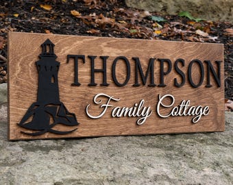 May include: A wooden sign with the text "Thompson Family Cottage" in white cursive lettering. A black silhouette of a lighthouse with waves is on the left side of the sign.