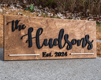 May include: A wooden sign with the text "The Hudsons Est. 2024" in black lettering. The sign has a brown wood grain finish and a white painted line.