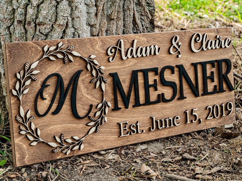 May include: A wooden sign with the text "Adam & Claire Mesner Est. June 15, 2019". The sign has a decorative wreath with a large "M" in the center.