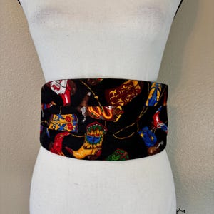 May include: A black fabric belt featuring a repeating pattern of colorful cowboy boots, purses, and other Western-themed imagery. The belt is wrapped around a white mannequin torso, showcasing its design and fit.