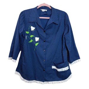 Può includere: Camicia blu navy con pois bianchi. La camicia presenta un ricamo floreale con fiori bianchi e foglie verdi. Ha un bordo in pizzo bianco sui polsini, sulla tasca e sull'orlo. La camicia ha maniche a tre quarti.