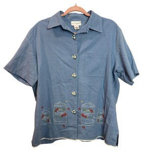 Puede incluir: Una camisa azul claro de manga corta con botes bordados con las etiquetas "Coffee" y "Sugar". La camisa tiene cuello, bolsillo en el pecho y botones con forma de tetera. El dobladillo tiene un ribete de encaje blanco.