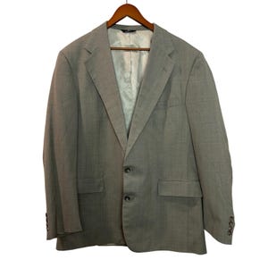 Peut inclure: Un blazer gris clair avec une fermeture à deux boutons, des revers crantés et deux poches avant. Le blazer est suspendu à un cintre en bois sur fond blanc. Les manches ont quatre boutons.