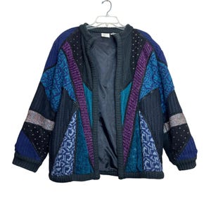 May include: A vintage patchwork cardigan in shades of blue, teal, purple, and black. The open-front jacket features a variety of textured fabrics and patterns, including ribbed knit, geometric designs, and dotted accents. The jacket is hanging on a white hanger.