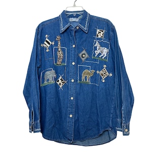 May include: A blue denim long-sleeve button-down shirt featuring embroidered patches of a giraffe, zebra, elephant, and camel. The shirt includes decorative stitching on the collar and cuffs, along with small animal print fabric squares.