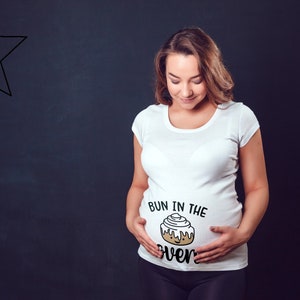 May include: A white t-shirt with the text "Bun in the oven" and a cartoon cinnamon bun graphic on the front. The shirt is being worn by a person with a pregnant belly.