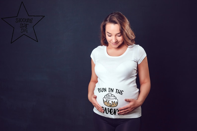 May include: A white t-shirt with the text "Bun in the oven" and a cartoon cinnamon bun graphic on the front. The shirt is being worn by a person with a pregnant belly.