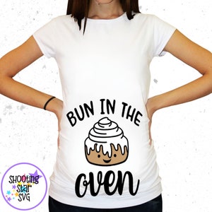 May include: White t-shirt with a black graphic that says "Bun in the oven" with a cartoon cinnamon bun illustration.