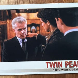 Könnte beinhalten: Eine Vintage-Sammelkarte mit einer Szene aus der Fernsehserie "Twin Peaks". Die Karte zeigt zwei Männer in einem schwach beleuchteten Raum, einer trägt einen Anzug und der andere eine dunkle Jacke. Die Karte trägt den Titel "Twin Peaks" und den Slogan "Drive with a dead girl".