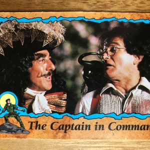 May include: A vintage trading card featuring a man dressed as Captain Hook from the movie "Hook" with a black beard and a red and gold costume. The card also features a man with brown hair and a white shirt, holding a black phone. The card has a blue border and the text "The Captain in Command".