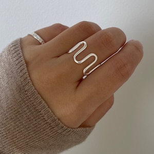 May include: A silver ring with a wavy, abstract design. The ring is worn on the middle finger of a hand.