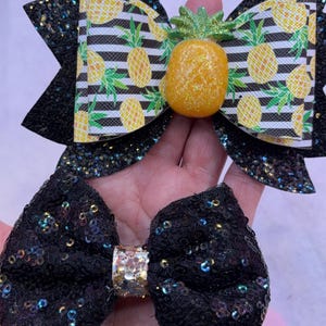 May include: Two hair bows, one with a black glitter fabric and the other with a black and white striped fabric with a pineapple print. The pineapple bow has a 3D pineapple accent.